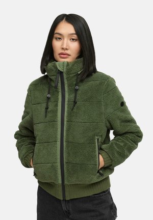 Femme avec des cheveux noirs lisses portant une veste en polaire verte zippée avec des cordons et les mains dans les poches, se tenant devant un fond blanc.