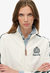 Hvid zip-up jakke med grønne detaljer og broderet logo. Bæres over en lyseblå og grøn stribet skjorte med høj krave.