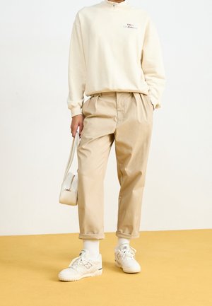 Person iført beige bukser, hvide New Balance sneakers, cremefarvet sweatshirt og holder en lille hvid håndtaske mod en ensfarvet lys baggrund.