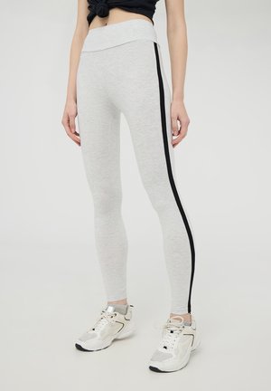 Leggings grigi chiaro con vita alta e striscia nera laterale, realizzati in tessuto morbido. Abbinati a sneakers bianche, creando un contrasto nel design.