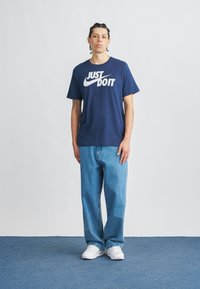 Μπλε ναυτικό μπλουζάκι με λευκό λογότυπο "JUST DO IT", συνδυασμένο με ανοιχτό μπλε τζιν και λευκά αθλητικά παπούτσια, φορεμένο από ένα άτομο που στέκεται μπροστά από ένα απλό φόντο.