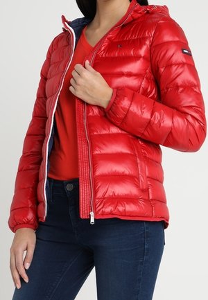 Femme portant une doudoune rouge avec capuche, la dézippant pour révéler un t-shirt rouge, associée à un jean bleu foncé.