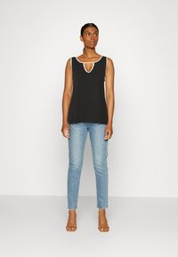 Top preto sem mangas com um corte em forma de buraco, apresentando detalhes brancos. Combinado com jeans azul claro e sandálias castanhas de tiras.