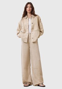 Chaqueta de lino beige con dos bolsillos frontales, botones plateados, combinada con pantalones de lino beige de pierna ancha y sandalias negras. Acabado de tejido texturizado.