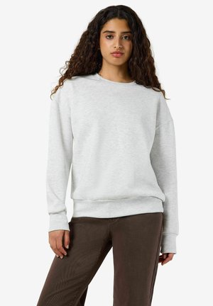 Femme aux longs cheveux bouclés portant un sweat-shirt gris clair et un pantalon marron foncé, debout avec une main le long du corps.