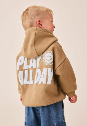Blond dziecko nosi beżową bluzę z kapturem z napisem „PLAY ALL DAY” i logo z uśmiechniętą buźką na plecach, w połączeniu z niebieskimi dżinsowymi szortami.