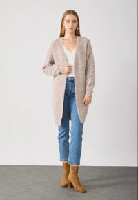 Cardigan longo bege feito de tecido de malha texturizada, usado sobre um top branco, combinado com jeans azul claro e botas de salto em tom de cáqui.