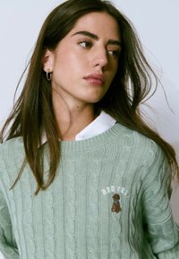 Joven mujer con cabello largo y oscuro lleva pendientes de aro de plata, una camisa blanca con cuello y un suéter de punto verde claro con un bordado de un perro pequeño.