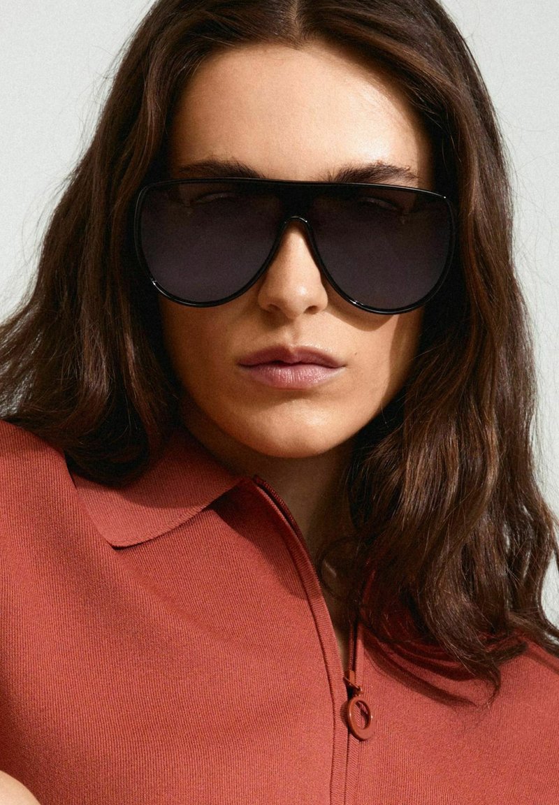 Oversized black sunglasses with gradient dark lenses, smooth plastic frame, worn with a rust-colored collared top featuring a zip closure.