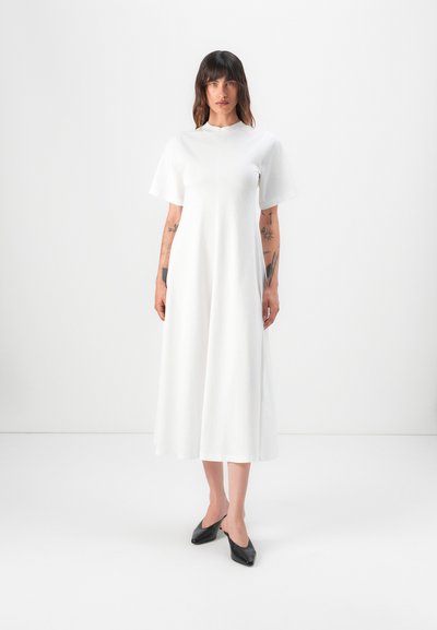 Witte jurk met korte mouwen, een hoge halslijn en een vloeiende, enkellange silhouet. Glad textuur, gedragen met zwarte, puntige schoenen.
