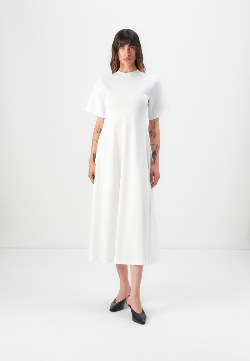 Weißes, kurzärmliges Kleid mit hohem Ausschnitt und fließender, knöchellanger Silhouette. Glatte Textur, getragen mit schwarzen, spitz zulaufenden Schuhen.
