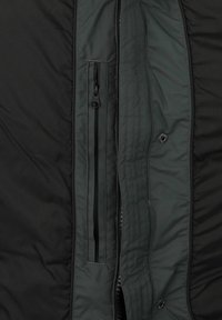 Interior de chaqueta aislante negra con un forro verde oscuro, que cuenta con un bolsillo vertical con cremallera y un detalle de botón a presión.