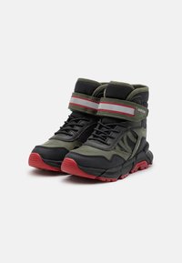Zapatillas altas en verde oscuro y negro con suela roja. Presentan un tobillo acolchado, correa de velcro y detalles de malla para la transpirabilidad.