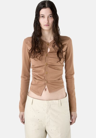 Femme aux longs cheveux foncés portant un haut marron froncé à manches longues boutonné et un pantalon taille haute beige clair avec de petites taches foncées.