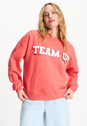 Vrouw met blond haar die een koraalkleurige sweater draagt met de tekst "TEAM UP" en lichtblauwe spijkerbroek, staand met handen bij de zakken tegen een witte achtergrond.