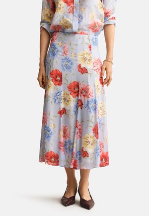Frau trägt einen hellblauen Midirock mit Blumenmuster in Rot, Gelb und Blau, kombiniert mit einer passenden Bluse mit Blumenmuster und dunklen spitzen Schuhen.