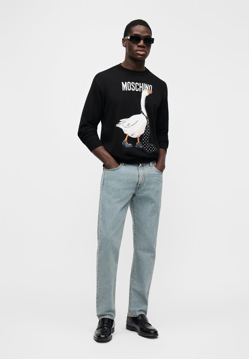 Schwarzer Pullover mit weißem Entenmotiv, blauen Details und Schriftzug "MOSCHINO". Hellblaue Jeans aus Denim und schwarze elegante Schuhe. Modell trägt Sonnenbrille.
