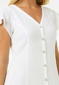 Robe blanche à manches courtes avec un décolleté en V, présentant un devant boutonné et un tissu texturé. Les boutons sont ronds et de couleur argentée.