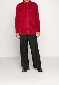 Camisa chaqueta de pana roja con botones en el frente, dos bolsillos en el pecho y mangas largas, combinada con pantalones de pierna ancha negros y zapatos negros.