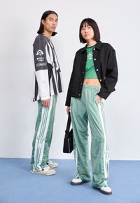 Dois modelos estão lado a lado. O homem veste uma camisola de manga longa às riscas preto e branco com um gráfico e bolsos; a mulher usa uma t-shirt cropped verde e uma jaqueta preta, com calças de treino verde claro com riscas e logótipos brancos.