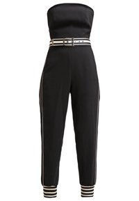 Combinaison noire sans bretelles avec bandes blanches sur les côtés, ceinture assortie rayée blanche et noire, et poignets de cheville rayés.