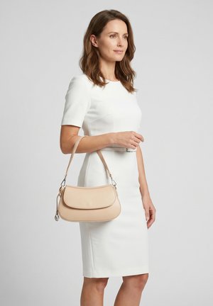 Femme portant une robe blanche ajustée, tenant un sac à main beige à l'épaule, debout devant un fond gris clair uni.