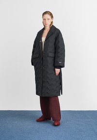 Μαύρο καπιτονέ μακρύ παλτό με γιακά, κλείσιμο με κουμπιά μπροστά και δύο πλαϊνές τσέπες. Φοριέται πάνω από μία κρέμα μπλούζα και μπορντό παντελόνι.