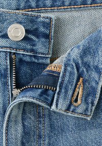 Gros plan d'un jean en denim bleu avec une fermeture éclair métallique partiellement fermée, une fermeture par bouton et des surpiqûres orange contrastantes sur les bords du tissu.