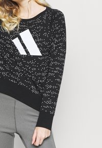 Sudadera negra con patrón geométrico blanco y un gran logo blanco en el frontal. Presenta una cintura ceñida y mangas largas.