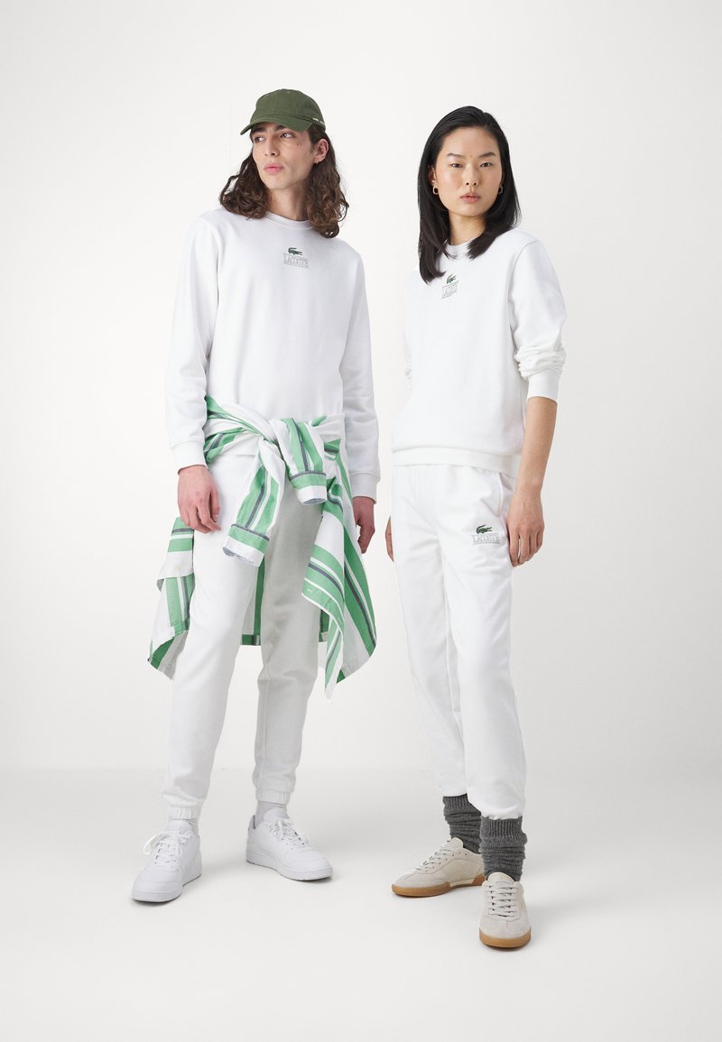 Due persone in felpe e pantaloni bianchi. Una indossa un cappello verde; l'altra ha una maglietta a righe verde e bianca legata in vita.