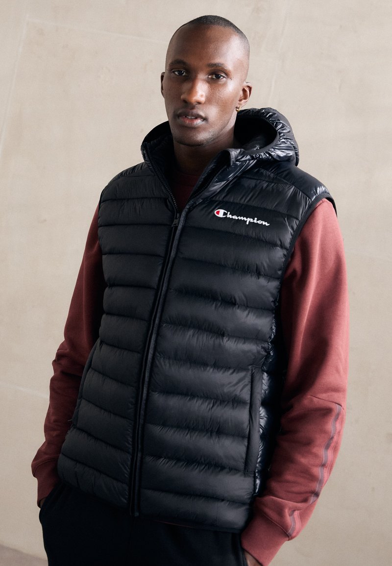 Chaleco de plumas negro con capucha, diseño acolchado, cierre de cremallera y logotipo bordado. Llevado sobre una sudadera burdeos. Textura suave.