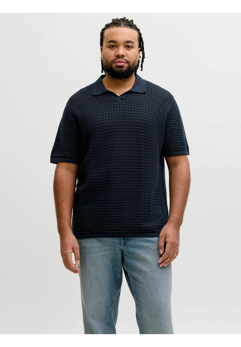 Homme aux cheveux tressés et à la barbe portant un polo noir texturé à manches courtes et un jean bleu clair, debout devant un fond uni.