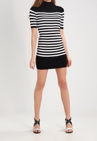 Musta-valkoraidallinen vartalonmyötäinen mekko, jossa lyhyet hihat, korkea kaulus ja istuva siluetti, yhdistetty mustavalkoisiin nauhallisiin korkokenkiin.