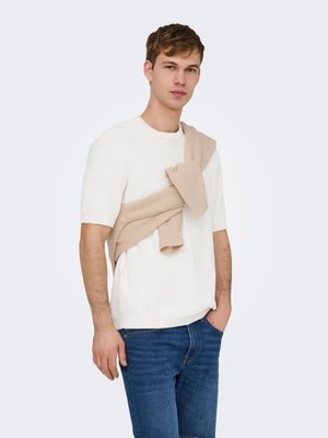 Jeune homme portant un pull blanc à manches courtes texturé avec un pull en maille beige posé sur les épaules, associé à un jean bleu, sur un fond uni.