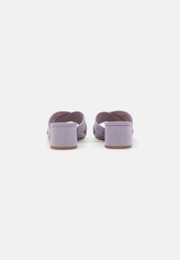 Mauve semišové muly na bloku s otevřenou zadní částí a křížovými popruhy. Hladká textura a hnědá gumová podrážka zajišťují stabilitu.