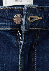 Jean en denim bleu foncé avec un bouton en métal argenté, des coutures jaunes et une fermeture éclair sur l'ouverture avant.