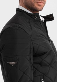Sort quiltet jakke med høj krave, sidelommer med lynlås og metallogo detalje på venstre ærme. Glat, letvægtsstof.