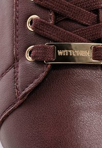 Sneaker in pelle bordeaux con superficie testurizzata, occhielli dorati e un'etichetta metallica con la scritta "WITTCHEN" attaccata ai lacci marroni.