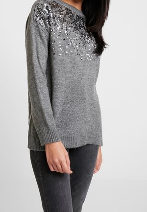 Femme portant un pull en tricot gris avec des sequins argentés et noirs sur le haut de la poitrine et l'épaule, associé à un jean gris foncé.