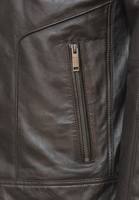 Nära bild av en brun läderjacka, med en dragkedjeficka med metallbeslag, slät textur och sömdetaljer.