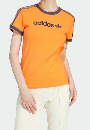 Osoba w pomarańczowej koszulce Adidas z czarnym logo i paskami na rękawach, w połączeniu z jasnymi spodniami.