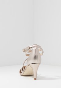 Metalické růžové zlaté sandály na vysokém podpatku s kroužkovým zapínáním, otevřenou špičkou a zúženým podpatkem. Mají hladkou texturu a minimalistický design.