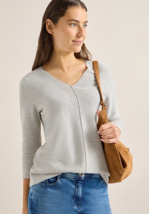 Femme aux longs cheveux bruns portant un pull gris clair à col en V et un jean bleu, tenant un sac à bandoulière en daim beige sur son épaule droite.