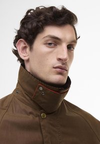 Brown jacket with a high collar, featuring red accent stitching and metal snaps. Textured fabric with a smooth finish and a fitted design.