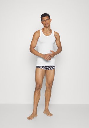 Débardeur blanc à fines bretelles, associé à un boxer noir à motifs présentant un design géométrique, modèle debout pieds nus.