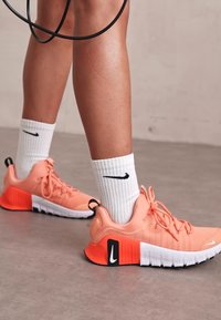 Svijetloružičaste atletske tenisice s bijelim detaljima i crnim Nike logom, uparene s bijelim rebrastim čarapama koje imaju crni logo, na sivoj podlozi.