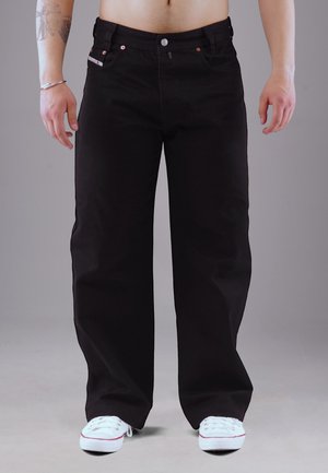 Man wearing loose black jeans and white sneakers, standing against a plain gray background, with tattoo and bracelet on left arm.