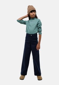 Haut à manches longues côtelé vert, col montant ; pantalon large marine ; bonnet en tricot marron ; chaussures souples beiges ; fond neutre.