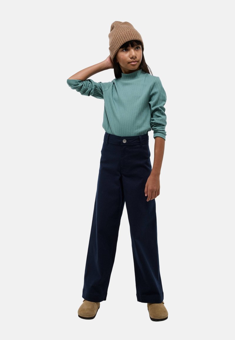 Haut à manches longues côtelé vert, col montant ; pantalon large marine ; bonnet en tricot marron ; chaussures souples beiges ; fond neutre.