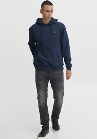 Marinblå bomullshoodie med en känguruficka, justerbar dragsko och en liten logotyp på bröstet, tillsammans med grå jeans och svarta sneakers.